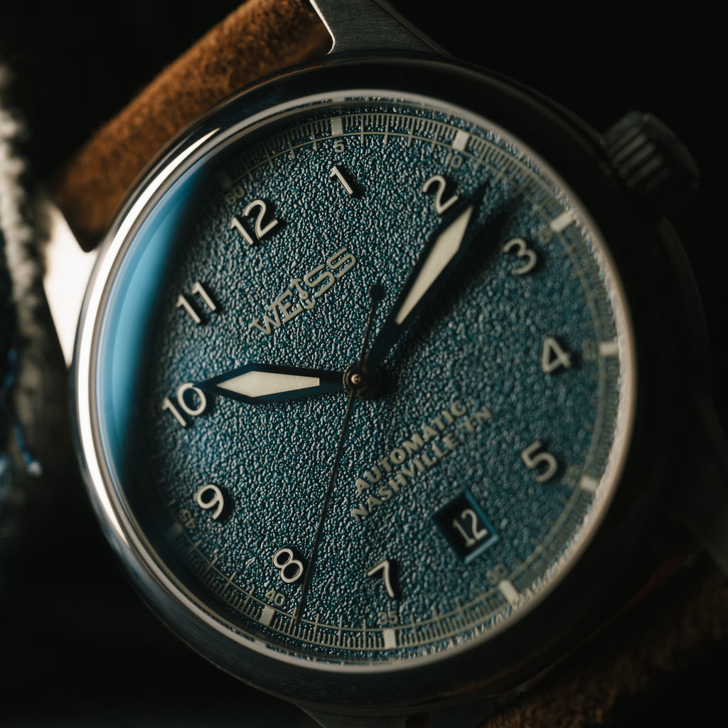 Close-up of a Weiss watch with a blue textured face and brown suede strap.